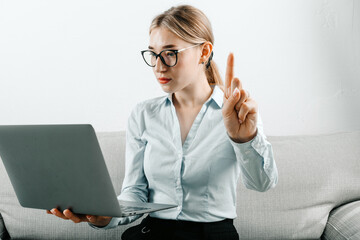 A young woman in glasses sitting on a couch, looking at her laptop screen with a shocked...