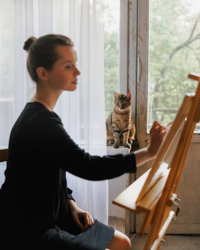 Woman is painting  picture while  cat watches her.