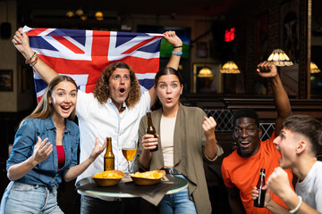 Multiracial United Kingdom sports fans, men and women, supporting their favourite team in bar, raising state flag and screaming chants together.