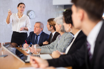 Successful smiling young business woman presenting new business strategy to partners in office sitting at table, switching presentation slides with clicker