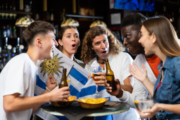 Multiracial Uruguay sports fans, men and women, supporting their favourite team in bar, raising state flag and screaming chants together.