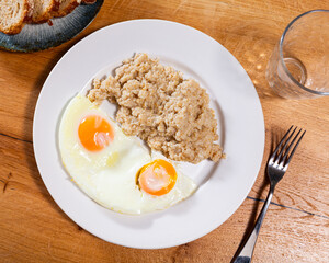 Oatmeal with two fried eggs on a wooden table view from above. Delicious breakfast option with whole grain porridge and farm eggs