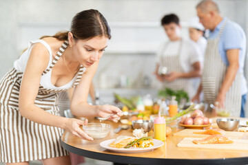 Positive young woman attendee of cooking course decorating meal made of salmon fish together with...