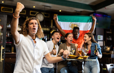 Cheerful multiracial soccer fans waving the flag of Iran while drinking beer and eating chips in sport bar