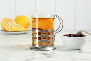 Glass cup of tea in metal holder with jam and lemon slices on white marble table, closeup