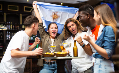 Group of emotional young adults Argentinean football fans cheering for favorite team together while watching match in sports bar