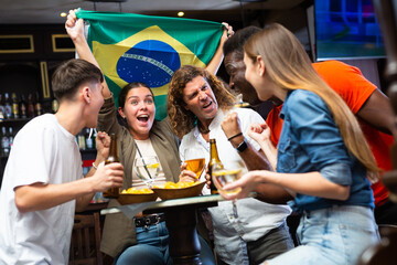 Happy international sport supporters holding up the flag of Brazil and drinking beer in the pub