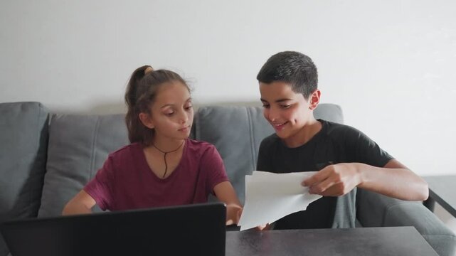 boy happily shows his handwritten work to girl seated beside him as she smiles while glancing at his paper then playfully slaps his head making him giggle and continue writing with dimples showing