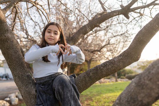 Young Girl Makes Heart Shape With Hands While Sitting in Tree Spring