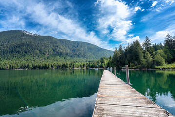 Idyllic view of a wooden pier in the lake with lush forested mountain in the background under wispy clouds