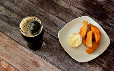 A tall glass of dark beer and a plate with a bunch of wheat croutons with sauce on a dark wooden table.