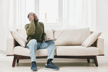 Obraz premium Frustrated young African man sitting on a couch with a laptop, expressing stress and anxiety about work or studies in a modern, bright living room environment