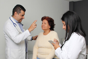 Fototapeta premium 80-year-old woman has her eyes, ears, and throat examined by a dark-skinned Latino doctor and his intern in the office as a checkup
