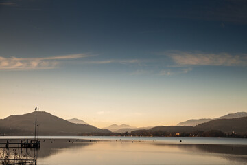Scenic view of Worthersee lake at dusk in Klagenfurt, Austria, capturing the tranquil reflection of mountains and sky during twilight.
