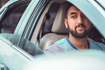 A bearded man sits inside a car, looking outside with a calm expression. The window is partially...