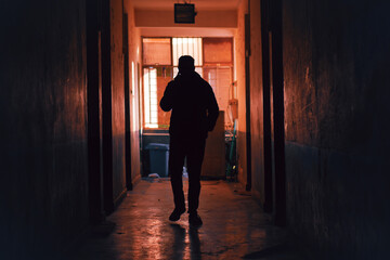 Silhouette of a person walking through a dimly lit, worn hallway toward a bright light at the end.