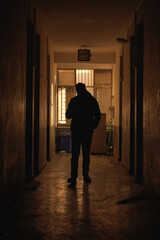 A worn hallway illuminated softly at the far end, where a silhouetted figure stands near a barred...