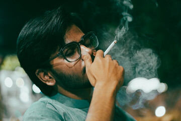 A young man with glasses holds a lit cigarette and smoking, smoke swirls around their hand, set...
