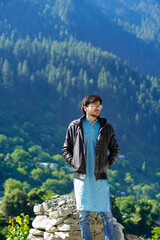 Naklejka premium Young traveler in a black jacket and blue jeans stands on rocks with hands in pockets, forested mountains provide a serene backdrop.