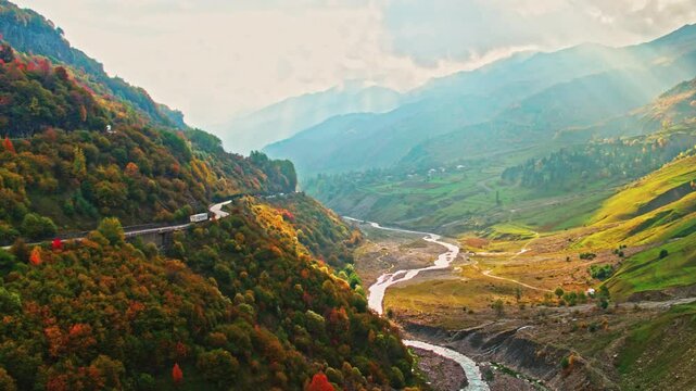 An aerial drone flight following a truck with RV trailer driving up a winding road through the Rocky Mountains of Georgia, Kazbegi-Gudauri during the colorful autumn season. River and big Mountains