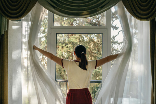 A middle-aged woman opens the curtains in a hotel in the morning.