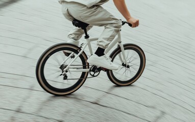 Fototapeta premium Person riding a white fixed-gear bicycle on a sloped, textured surface. The cyclist wears light beige pants and white sneakers. The image is taken from a low angle and has a blurry background