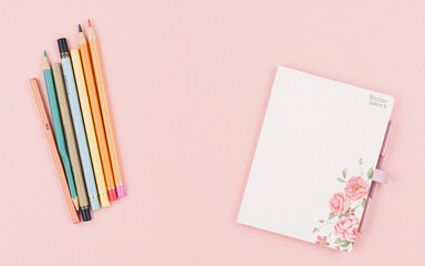 Stationery items including colored pencils and a floral notebook are arranged on a light pink surface from a top-down perspective.