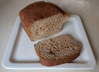 Rustic whole wheat bread loaf with a sliced piece on a clean white plate, photographed in natural light with clear texture detail.