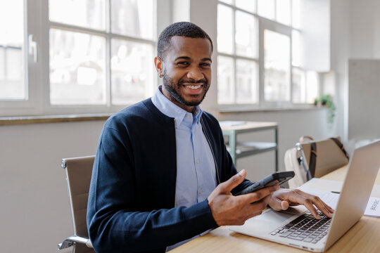 Smiling formal businessman using laptop and smartphone in office