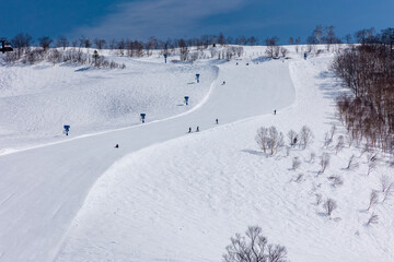Snowboarders descending a wide, steep ski slope on bright sunny day