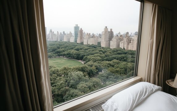Fototapeta Serene view of Central Park from a hotel room window. Lush green trees, city buildings, and a partially visible bed are seen in the image. Natural light illuminates the scene, giving a calm and