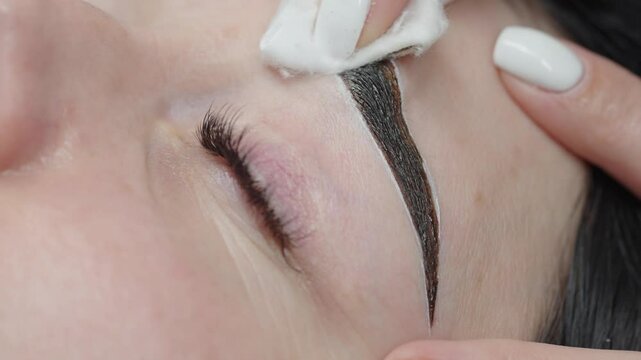 A beautician gently wipes excess eyebrow dye with a cotton pad during a tinting procedure, revealing a clean and defined brow shape.
