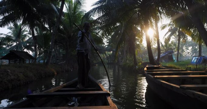 Silhouette of canoe driver gently pushing through Kerala backwaters at sunrise. Traditional boat glides under palm trees in warm light, evoking peaceful travel and cultural authenticity.