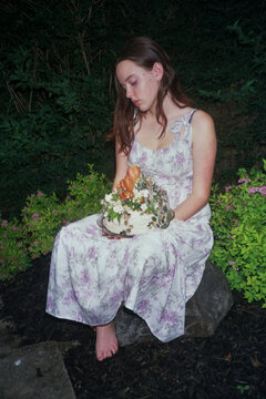 Young Woman in Floral Dress holds a Decorated Cake Outside