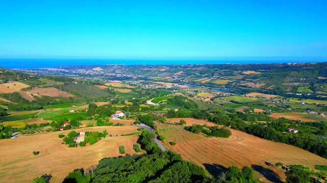 Smooth aerial push-in shot from Moresco (FM), brown fields punctuated by green trees, thickets, houses, buildings, roads, checkered hillscapes, swathes of blue Adriatic Sea, clear gradient azure sky