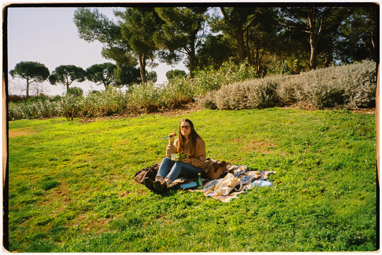 Film wide shot of woman enjoying food sitted on a blanket on the grass