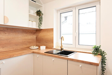 Small kitchen with white furniture, wooden cabinets and kitchen sink with faucet near the window. Modern interior of room in new apartment and wood on the wall. Real estate.	