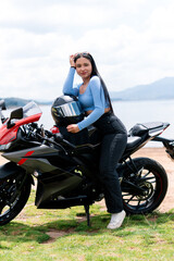 Young woman biker posing leaning on her motorcycle with her helmet resting on the seat, near a lake and mountains in the background
