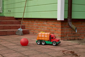 Children's toy truck and red ball placed on a brick patio near a broom and green siding. Scene depicts an everyday outdoor playtime setting with simple toys.