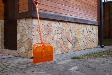 Bright orange plastic snow shovel rests against a rustic stone wall of a wooden house. The scene...