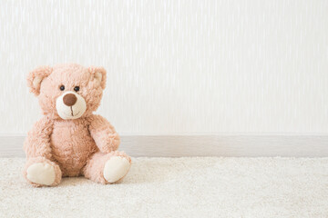 Smiling brown soft teddy bear sitting on soft white home carpet at light gray wall background. Empty place for text, quote or sayings. Closeup. Front view.