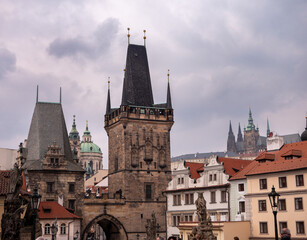 View of the Lesser Town Bridge Towers on Charles Bridge in Prague, with St. Vitus Cathedral and baroque domes in the background under a cloudy sky.