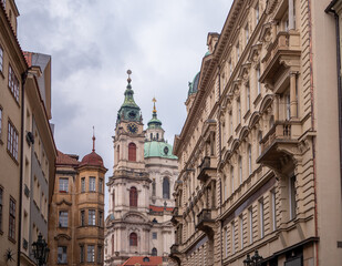 Fototapeta premium St. Nicholas Church in Prague’s Lesser Town, featuring stunning baroque architecture, green domes, and clock tower framed by surrounding historic buildings.