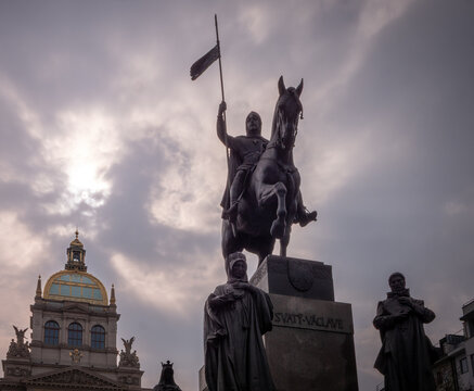 Silhouette of the Saint Wenceslas statue on Wenceslas Square in Prague with the National Museum in the background under a dramatic cloudy sky.
