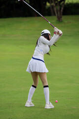 Obraz premium Asian woman happy and fun while playing golf on the golf course. Golfer hitting golf shot with driver club on course. Beautiful woman smiling and holding a golf club while standing on green lawn.