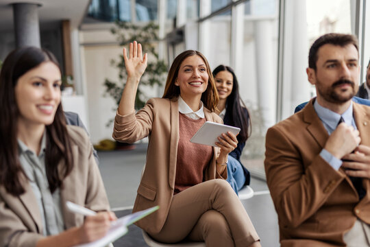 Young professional businesswoman sitting at training room with her colleagues and holding her hand raised.