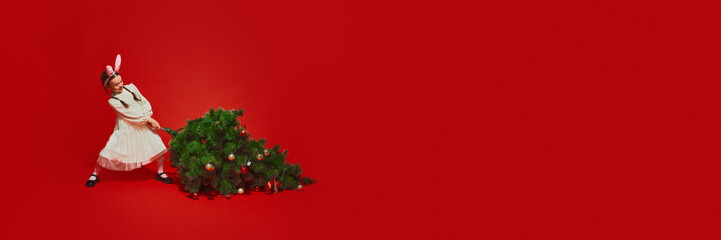 Young girl, child in festive white dress carrying decorated Christmas tree against red studio...
