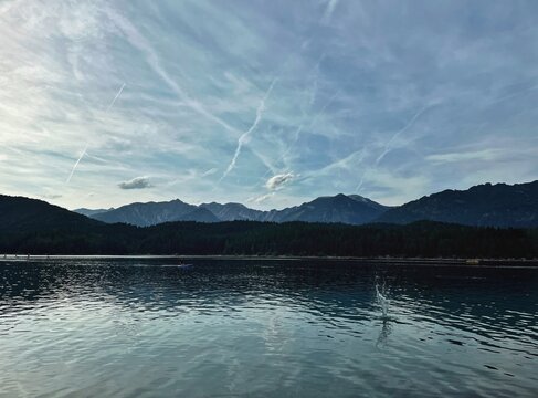 Wide view of a serene lake surrounded by mountains under a cloudy sky at sunset with jets leaving trails in the atmosphere