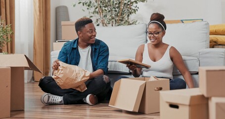 In the living room of their new house, a joyful couple sits among cardboard boxes. They unwrap a photo frame, a delicate vase, and treasured trinkets from paper.