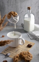 Oatmeal cookie dripping milk above a ceramic mug with splash. Styled breakfast scene with milk bottle, cookies, spices and sheet music. Perfect for food styling, slow living and cozy kitchen content.